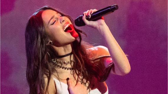 A close-up of Olivia Rodrigo who holds a microphone in the air as she sings