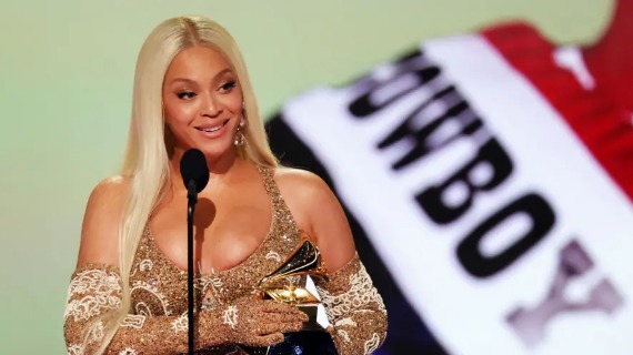 Beyonce has long, platinum-blonde straight hair. She is wearing a golden dress with elbow length gloves and is speaking in front of a microphone while holding a grammy award. 