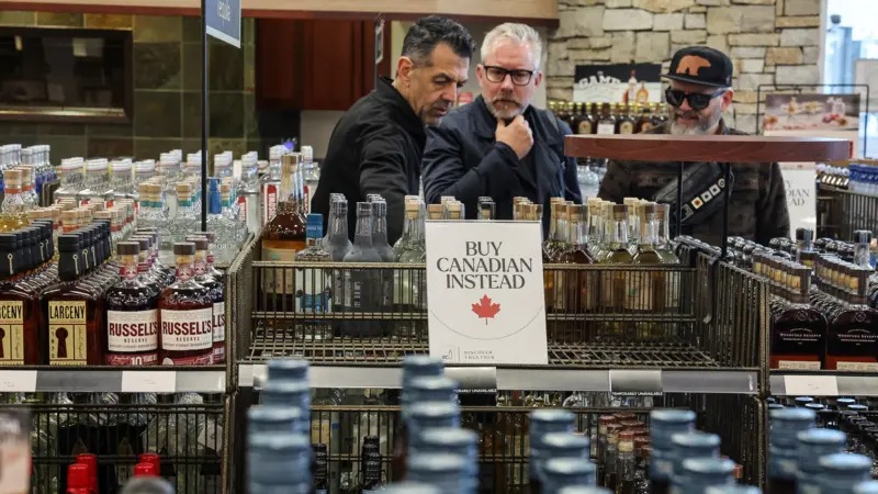 A sign reading 'Buy Canadian Instead' featuring a red maple leaf, is displayed on an empty shelf at a liquor store - the shelves around it are fully stacked.