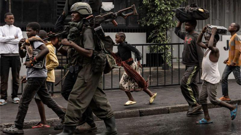 Civilians carry bags on their heads and run past an M twenty three rebel carrying a light machinegun, and wearing green combat fatigues and a helmet