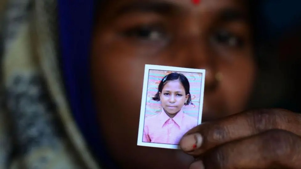 A woman holds up a photo of a young girl, who has short black hair and is wearing a pink shirt.