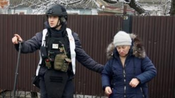 A Ukrainian rescuer in a military style helmet and tactical gear escorts an elderly lady in a winter coat and hat