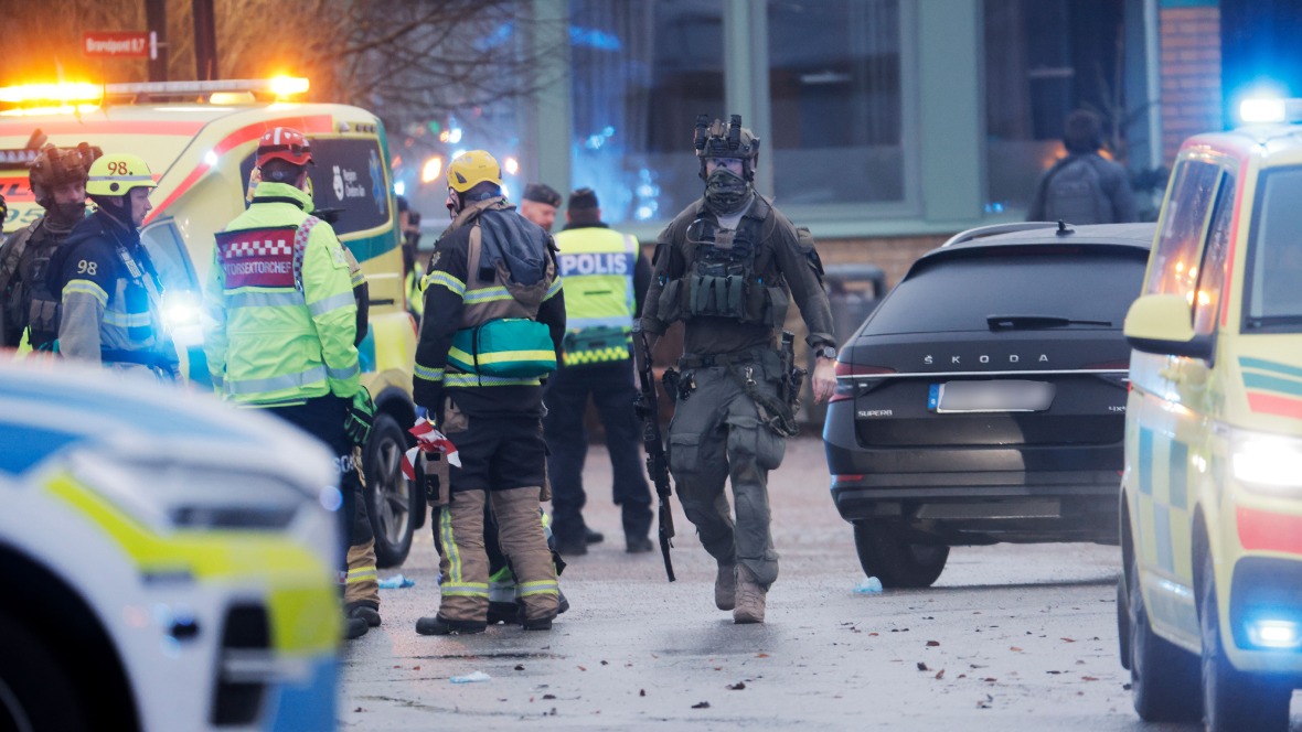 A police officer in full tactical gear holds a rifle as he walks past emergency services vehicles