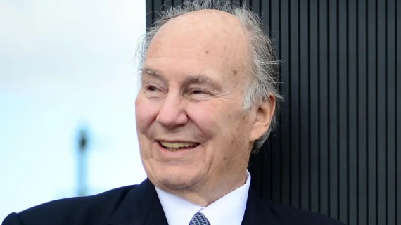 Prince Karim Aga Khan has white hair and brown eyes. He is pictured smiling, wearing a dark suit, white shirt and blue spotted tie.