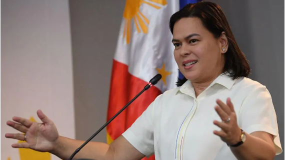 Sarah Duterte has short brown hair. She is speaking in front of a microphone wearing a white shirt. The Philippines flag stands in the background.