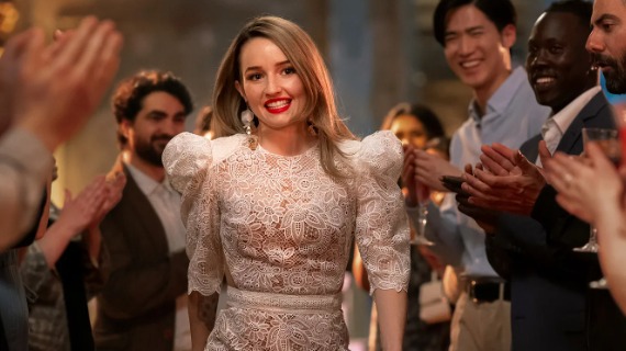 A screenshot from Apple Cider Vinegar shows Kaitlyn Dever in character as Belle Gibson, wearing a lace white dress and smiling as she walks through a crowd of people who are applauding her.