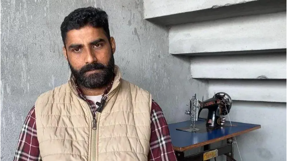Jaspal Singh has black hair and a black beard. He is wearing a plaid shirt and a beige gilet. A sewing machine is laid on a table in the background.