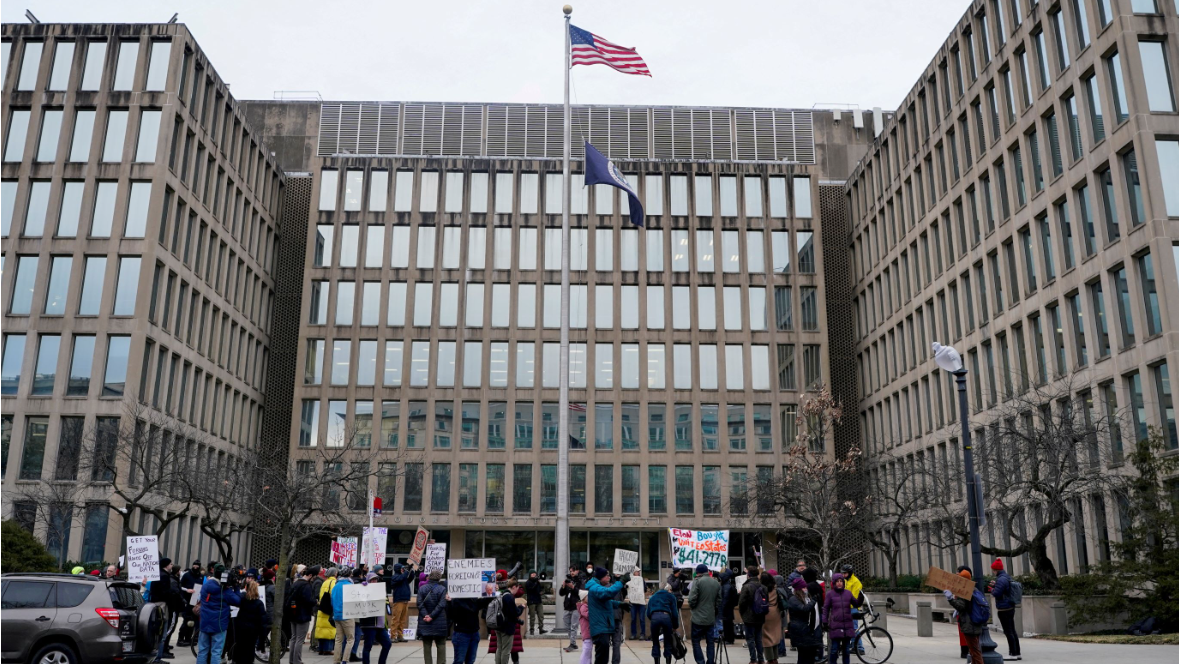 Tens of thousands of federal workers accept Trump resignation offer The US flag and the US flag bearingthe presidential seal fy on a mast in front of an imposing looking building. Dozens of people are demonstrating in front of the building, holding up signs.