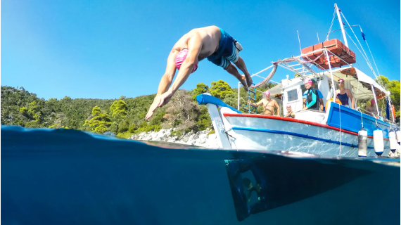 A man in a pink swimming cap and blue swimming trunks dives headfirst into water from a boat where other people in similar swimming gear are standing.