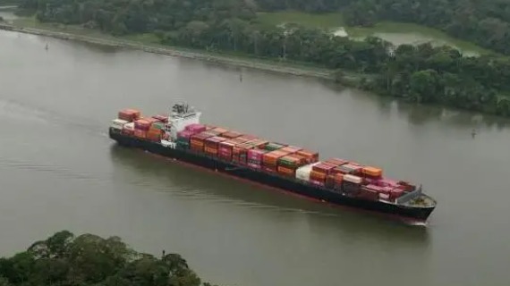 A cargo ship stacked with brigh orange and red containers floats through the Panama Canal, which features green vegetation along its banks.