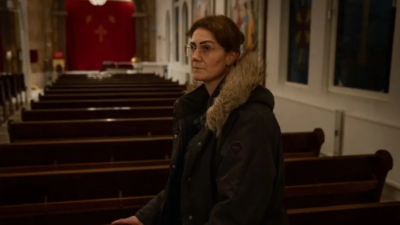 Shooting survivor Nour Afram has brown hair and brown eyes. She is wearing glasses and a black winter jacket. She leans a hand on the seats of the Orthodox church she is a member of.