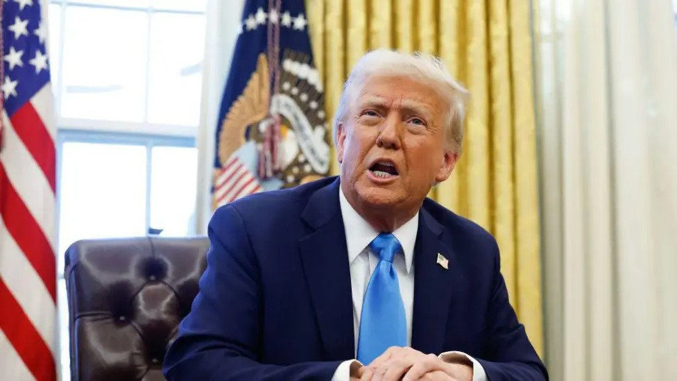 Donald Trump has white-blond hair and blue eyes. He is wearing a blue suit, white shirt and a light blue tie. He is sat in the Oval Office, with the US flag and a banner featuring the presidential seal in the background.