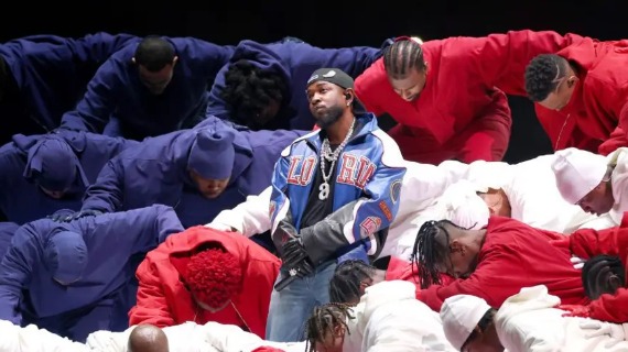 Kenrick Lamar performs in a baseball hat, jeans, and a varsitiy jacket. Behind him, dancers wearing blue, red, and white overalls are arranged into rows resembling the US flag. 