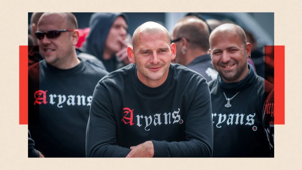 Men with their head shaven wear black crewneck jumpers with the word 'Aryans' imprinted on it in gothic font. The letter A is in red while the rest of the word is in white.