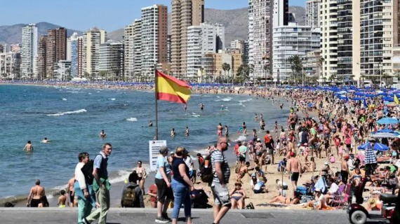 A Spanish flag flies on a mast perched on a beacj packed wtih people, tall buildings rising in the background.