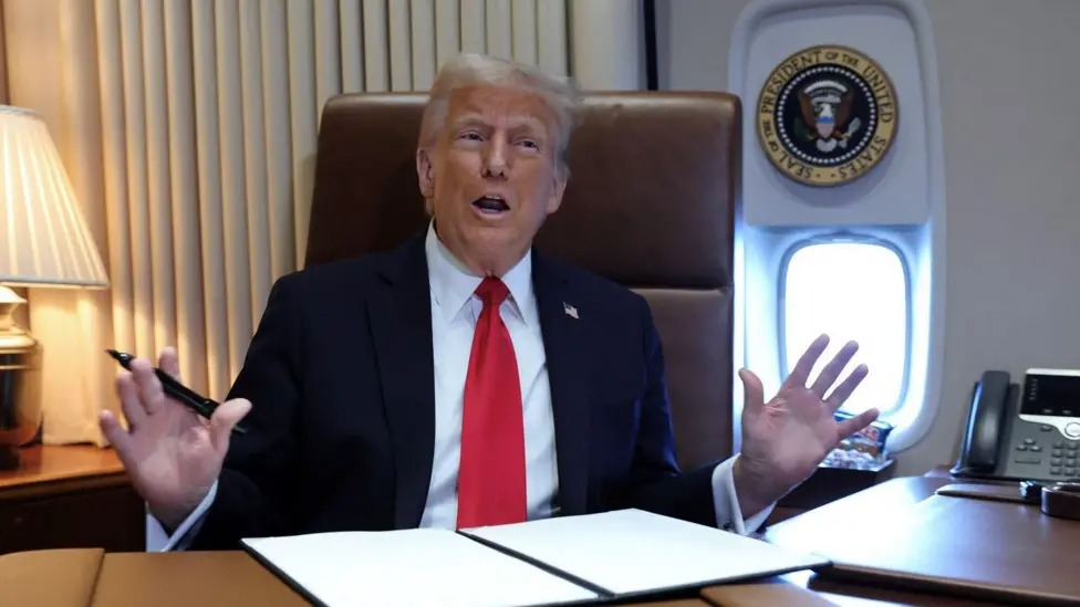 Donald Trump sits on a leather chair at a desk on Air Force One, the presidential seal displayed above an airplane window behind him. He is holding a pen and has a folder containing papers opened in front of him.