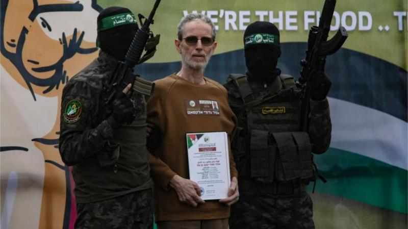 An Israeli, Ohad Ben Ami wears a brown t-shirt and holds a certificate of release as he is flanked by two masked Hamas militants in green fatigues holding assault rifles