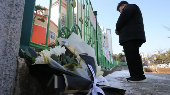 A bunch of white flowers are laid on the pavement against the green gate of a school in South Korea. A mourner can be seen in the background.