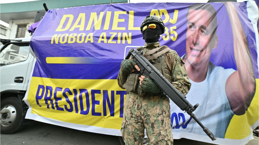 A masked soldier in military fatigues holds his weapon as he stands in front of a campaign poster for Daniel Noboa, which shows him waving and smiling.