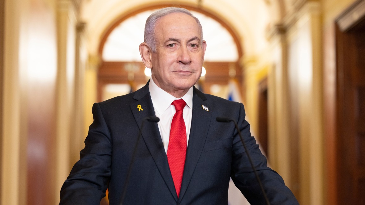 Benjamin Netanyahu wearing a dark blue suit and red tie standing at a podium and looking at the camera.