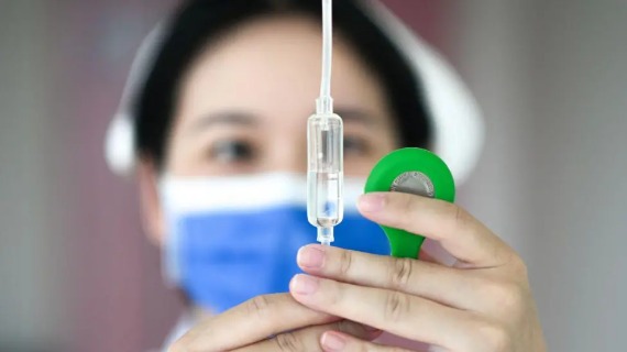 A nurse with her hair covered and wearing a face mask checks the flow of a drip