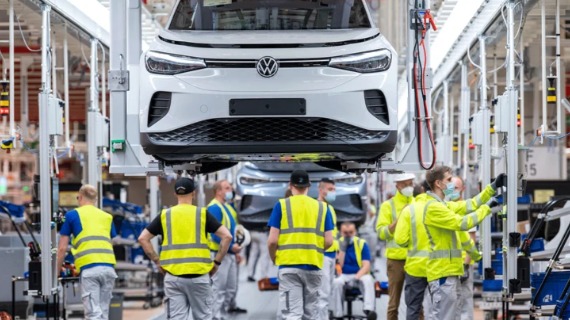 A white Volkswagen car is suspended on a platform inside a factory where a group of workers are wearing high-vis jackets and gilets.