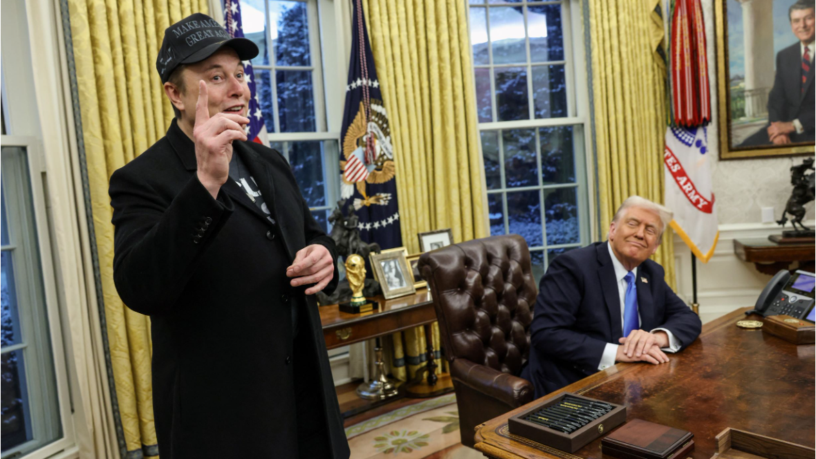 Elon Musk points his right index finger upwards. He is wearing a black baseball hat, a black tshirt and a black coat. Sitting at the Resolute Desk, Donald Trump smiles as he looks in his direction.