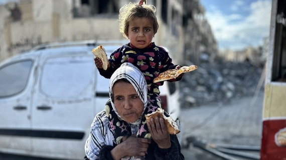 A child holds bread as she sits on the sholders of a woman in a rubble-strewn street in Gaza